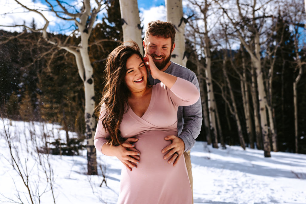 Parents to be laughing and embracing  pregnancy in the snowy mountains of colorado 