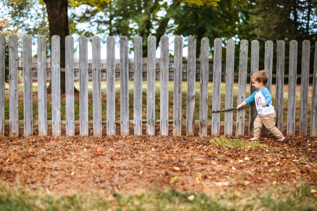 Two year old boy playing with a stick along a fence, at home family photos session in High Points, North Carolina 