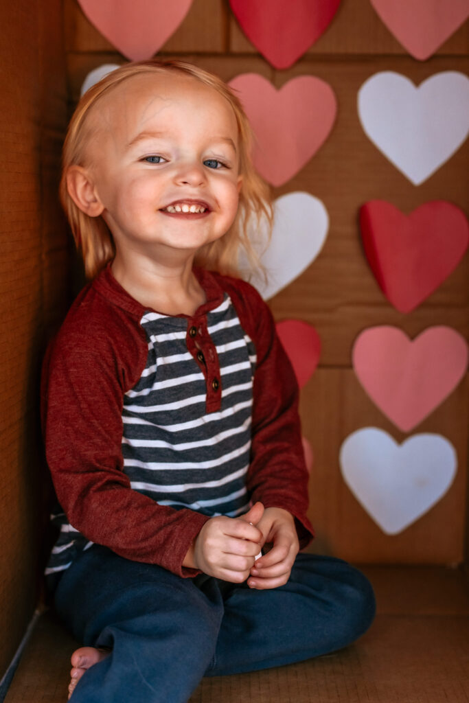 Toddler boy in a cardboard box themed with Colored Hearts for Valentines Day 